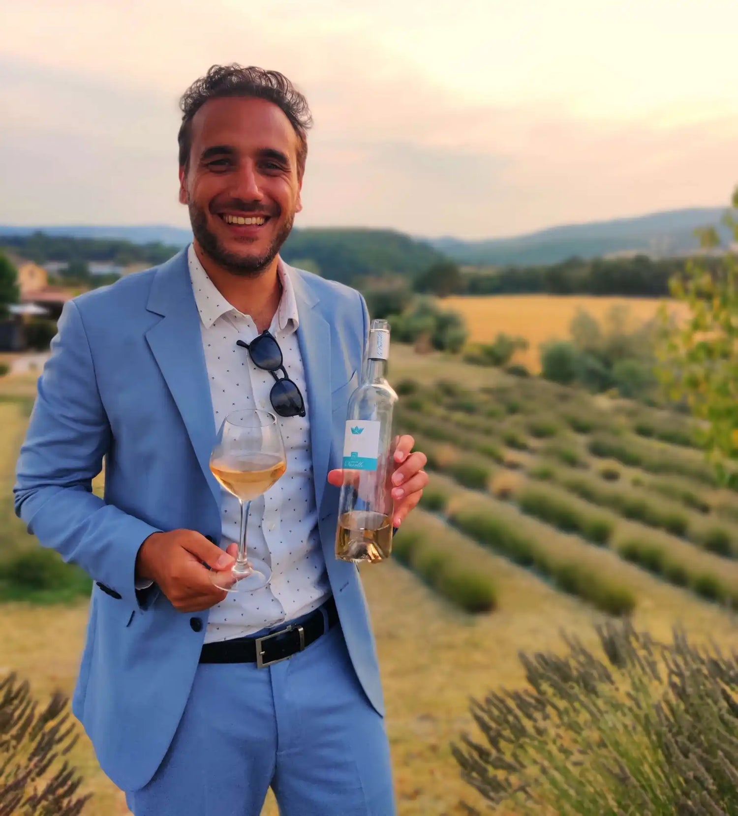 Un homme souriant en costume bleu clair tenant un verre de vin rosé et une bouteille de vin blanc.